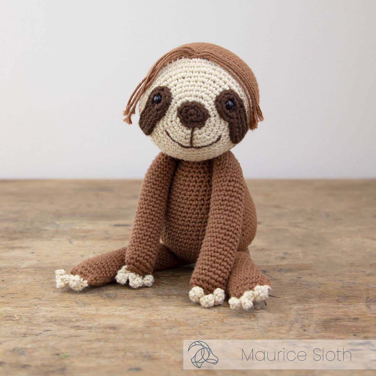 Hand-crocheted sloth toy sitting upright on a wooden surface, made in brown and cream yarn with long arms and a smiling face.