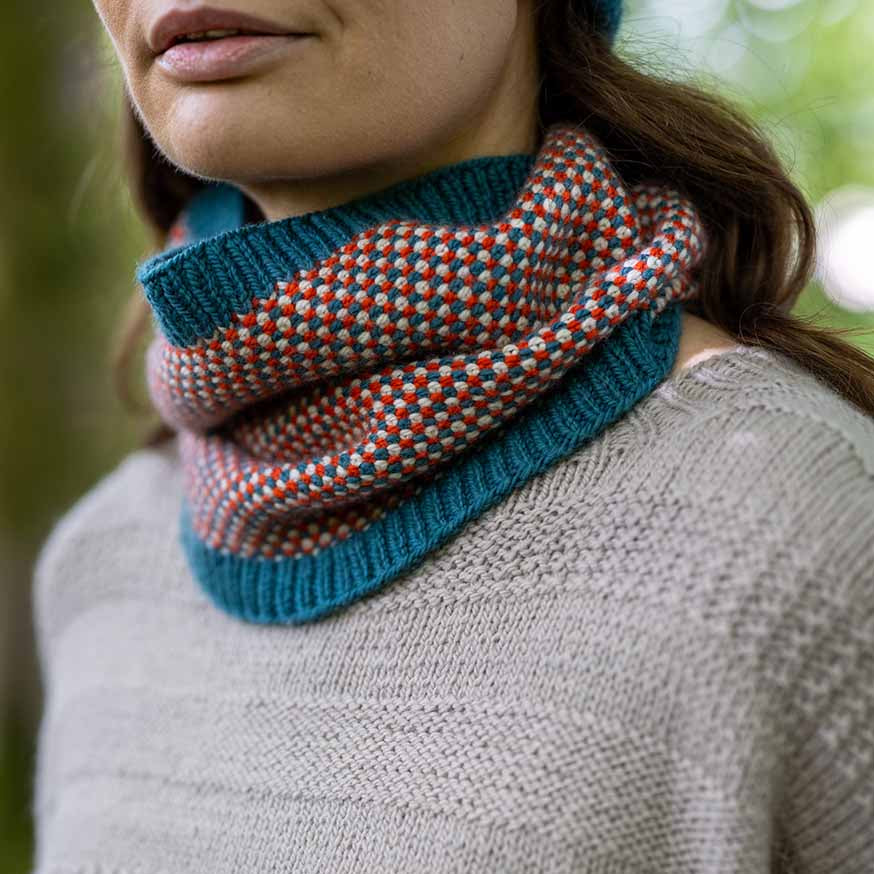 A close-up of a person wearing a knitted cowl neck warmer with a teal blue base and orange-red and light blue speckled pattern, paired with a beige knit sweater.
