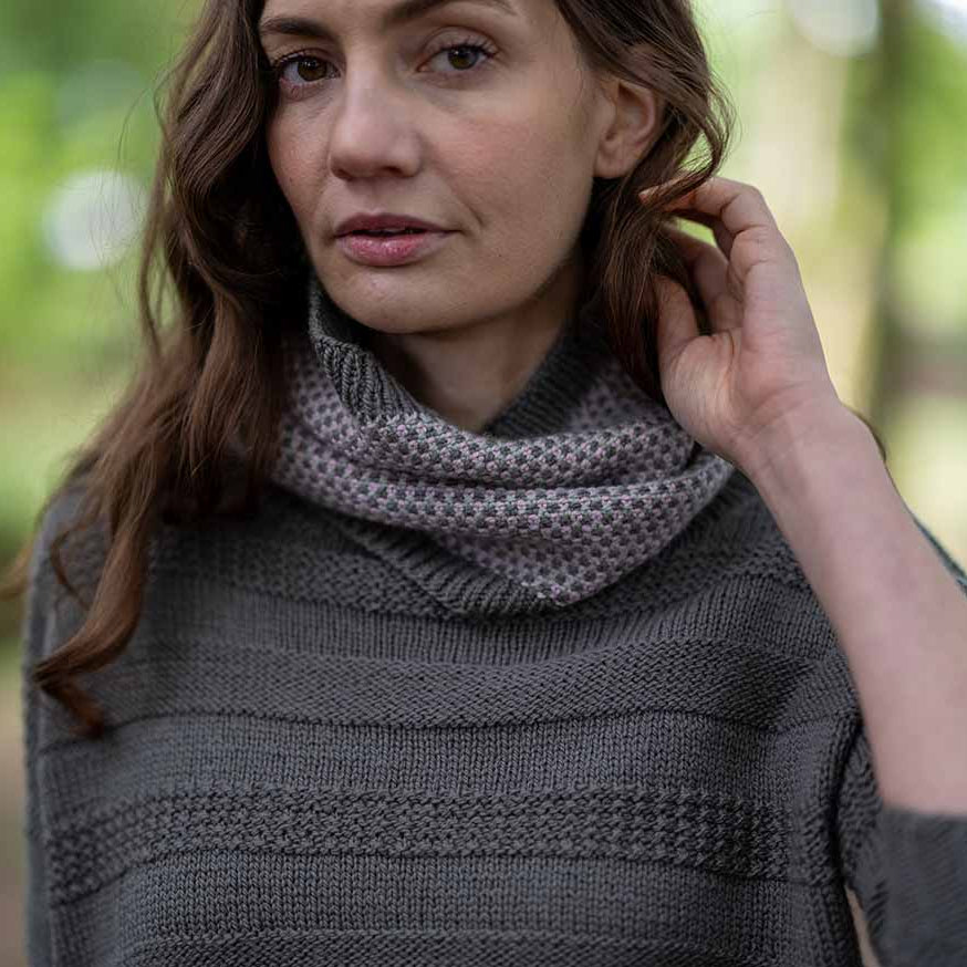 A woman wearing a dark gray knit sweater and a textured gray cowl neck scarf, photographed outdoors with a blurred forest background.