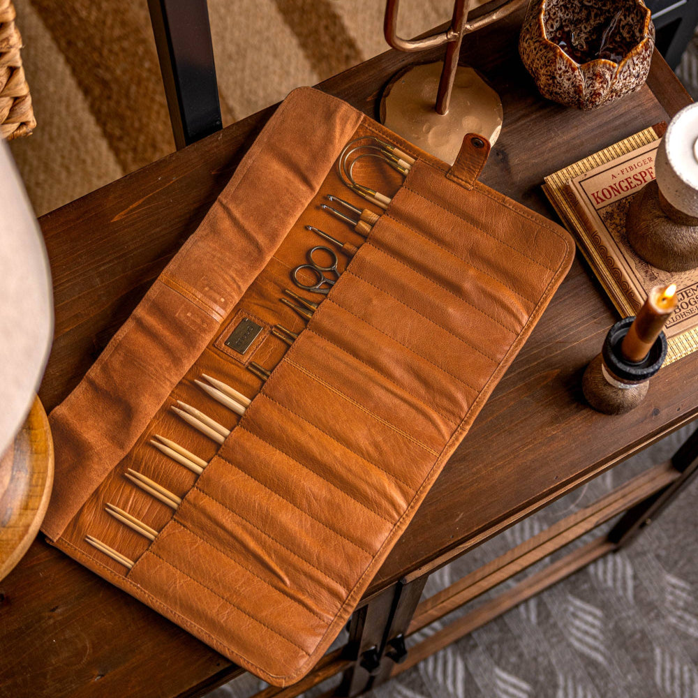 A brown genuine leather knitting needle case with multiple compartments, closed with a button, displayed on a wooden surface.