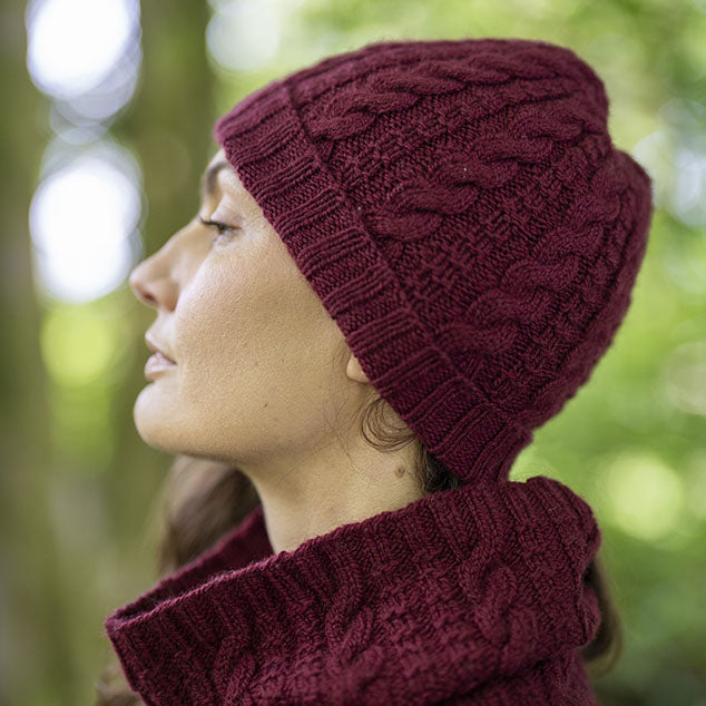 A woman in profile wearing a deep burgundy Darvel cable-knit beanie and matching  Darvel knit neck warmer, photographed outdoors against a soft green bokeh background.