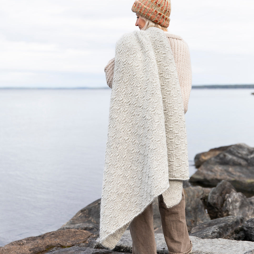 Person wrapped in a large off white blanket standing on rocks by the sea.