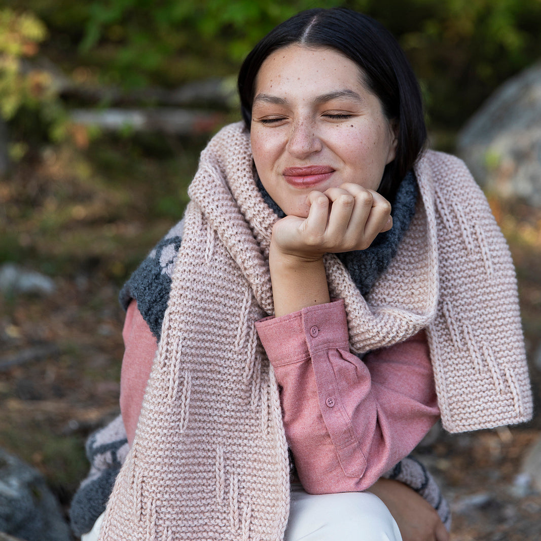 Woman wearing a pink shirt and beige scarf sitting outdoors with greenery in the background