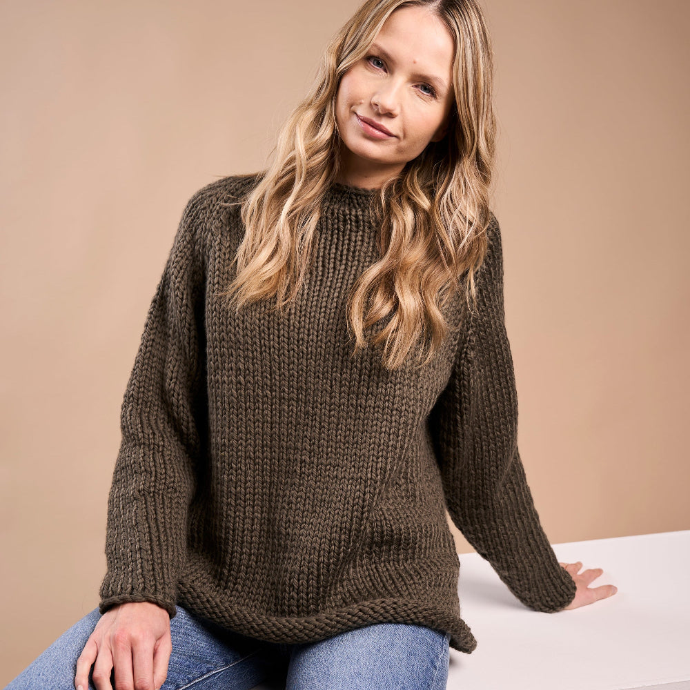 Woman wearing the Mae Sweater knitted in brown Rowan Big Wool she is also wearing blue jeans and sitting on a white stool against a beige background